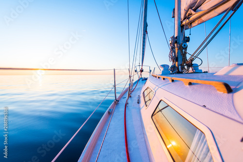 The calm water. White sloop rigged yacht sailing at sunset. A view from the deck to the bow, mast and the sails. Baltic Sea, Latvia photo