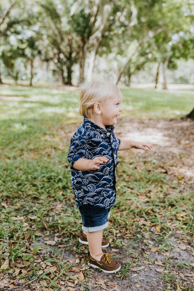 Adorably Happy and Cute Little Caucasian Toddler Baby Boy with Long Blond Hair Laughing, Playing, and Running Outside in Green Nature at the Park of Big Mature Oak and Moss Trees