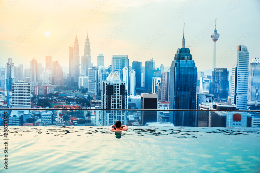 Naklejka premium Azjatycki biznesmen zrelaksować się w basenie na dachu za pięknym widokiem na miasto Kuala Lumpur w sunrise niebo, Malezja