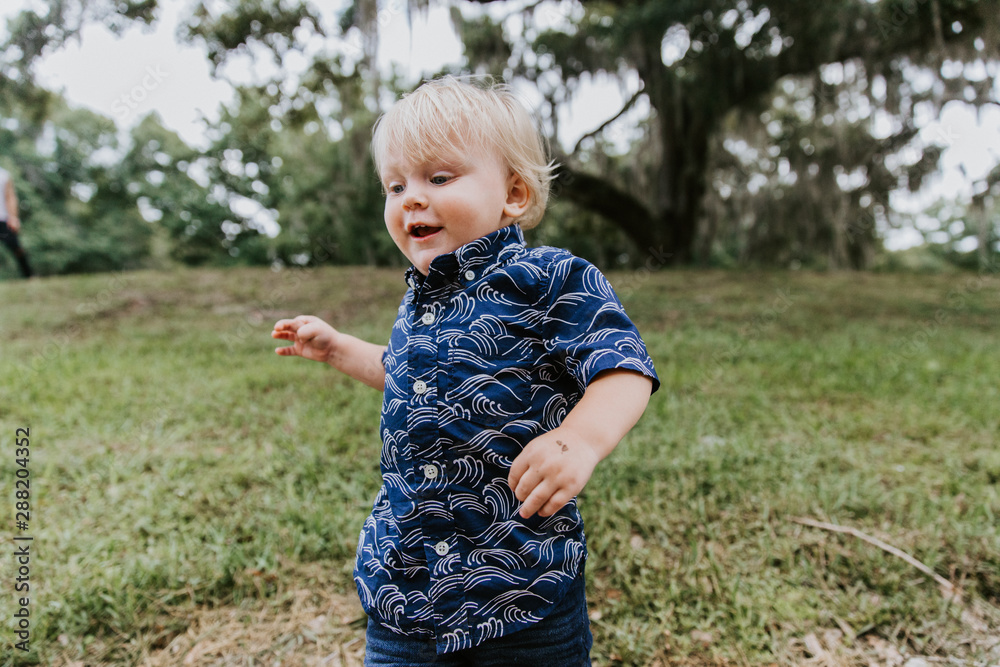 Adorably Happy and Cute Little Caucasian Toddler Baby Boy with Long Blond Hair Laughing, Playing, and Running Outside in Green Nature at the Park of Big Mature Oak and Moss Trees Adorably Happy and Cute Little Caucasian Toddler Baby Boy with Long Blond Hair Laughing, Playing, and Running Outside in Green Nature at the Park of Big Mature Oak and Moss Trees