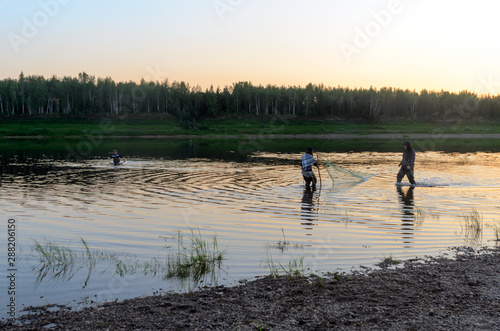 Two men Yakuts and graceful girl in a shawl to go in wading boots with fish net on the wildlife in the river Vilyuy in a forest traditionally catching local fish tugunok. photo