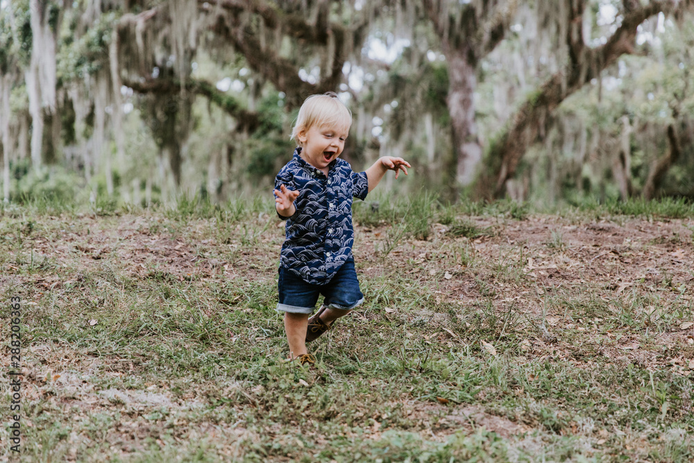 Adorably Happy and Cute Little Caucasian Toddler Baby Boy with Long Blond Hair Laughing, Playing, and Running Outside in Green Nature at the Park of Big Mature Oak and Moss Trees Adorably Happy and Cute Little Caucasian Toddler Baby Boy with Long Blond Hair Laughing, Playing, and Running Outside in Green Nature at the Park of Big Mature Oak and Moss Trees