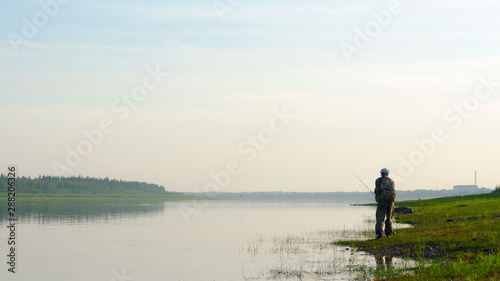Yakut Asian girl-tourist fisherman with a backpack and a hat to fish with a spinning in the river Vilyuy in the haze at sunset in the wild North of Russia on the background of the village Suntar.