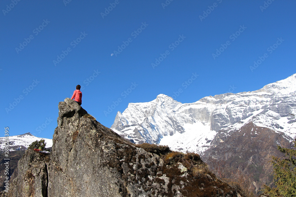 A woman with short hair, dressed in a red sweatshirt, sits on a rock in Nepal with Kongde Ri mountain in the background. Nature, people, outdoors, hiking, travel and tourism concept.