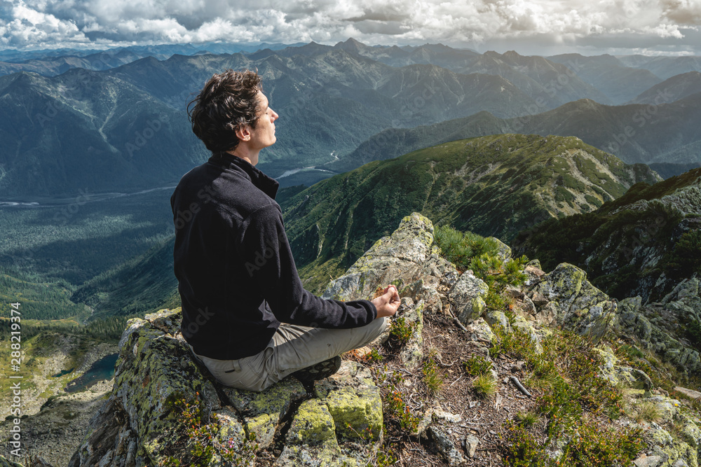 Fototapeta premium Man hiker sitting in a yoga pose at the peak of the mountain in the summer. Meditation after a long climb on a mountain