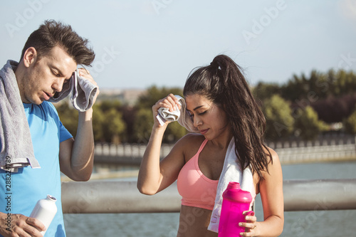 Interracial couple, Latina and Caucasian drink from a bottle of water with a towel around their neck after outdoor sports