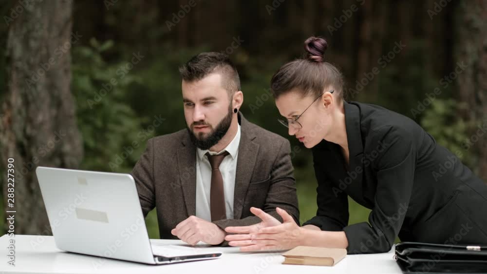 Young attractive male businessman in a brown suit and a woman in a jacket and glasses discussing work processes behind a laptop. Remote team work in the fresh air. Green plants around. Young attractive male businessman in a brown suit and a woman in a jacket and glasses discussing work processes behind a laptop. Remote team work in the fresh air. Green plants around.