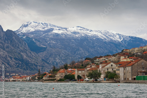Winter Mediterranean landscape. Small town at foot of mountains on cloudy day. Montenegro, Adriatic Sea, Bay of Kotor, view of Prcanj town and of snow-capped mountain of Lovcen