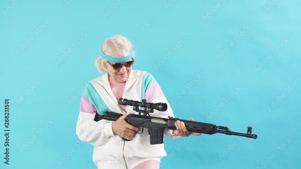 Experienced female agent working undercover of a grandmother-dandelion is pointing a rifle at camera, standing isolated on blue background. Poster. Blue background.