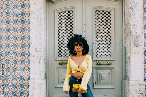 Trendy African American woman in yellow crop top and jeans standing by door and looking at camera photo