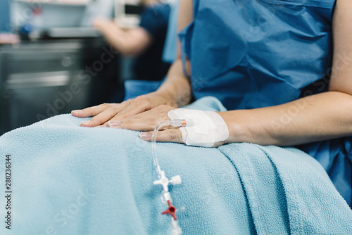 Crop female patient sitting with covered legs and intravenous fluid needle in hand before surgery in operating room photo