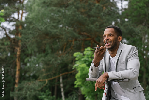 Elegant African American businessman recording voice message on mobile phone while riding on electrical scooter in park photo