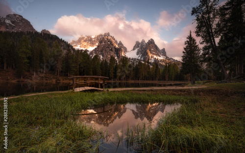 Pacifying landscape of dense forest along which path going with small bridge next to round pond spreading out on lush green lawn reflecting sky and mountains in Dolomites Itlay photo