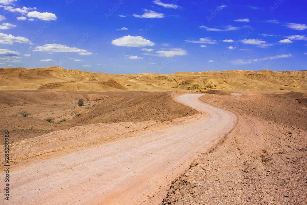 wasteland desert lonely curved trail through sand dunes to horizon with mountains hills and blue sky white clouds background 