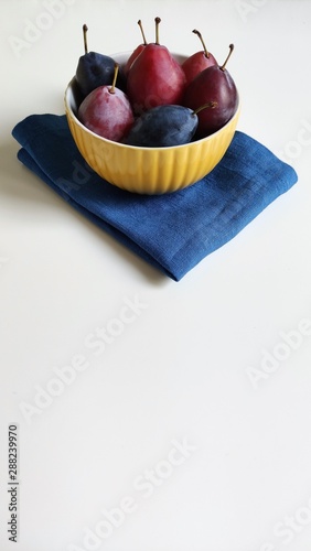 A yellow ceramic bowl with red and blue ripe plums on a white table against a washed-out gray wall. The plate is on a blue linen napkin. Side view, food photography.