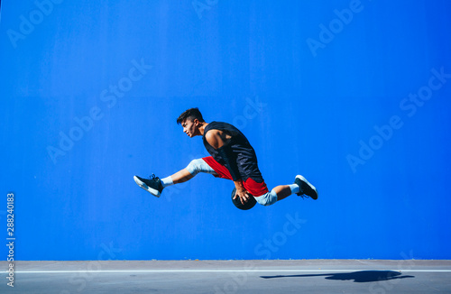 Side view of a young man holding a basket ball jumping in front of a blue wall.