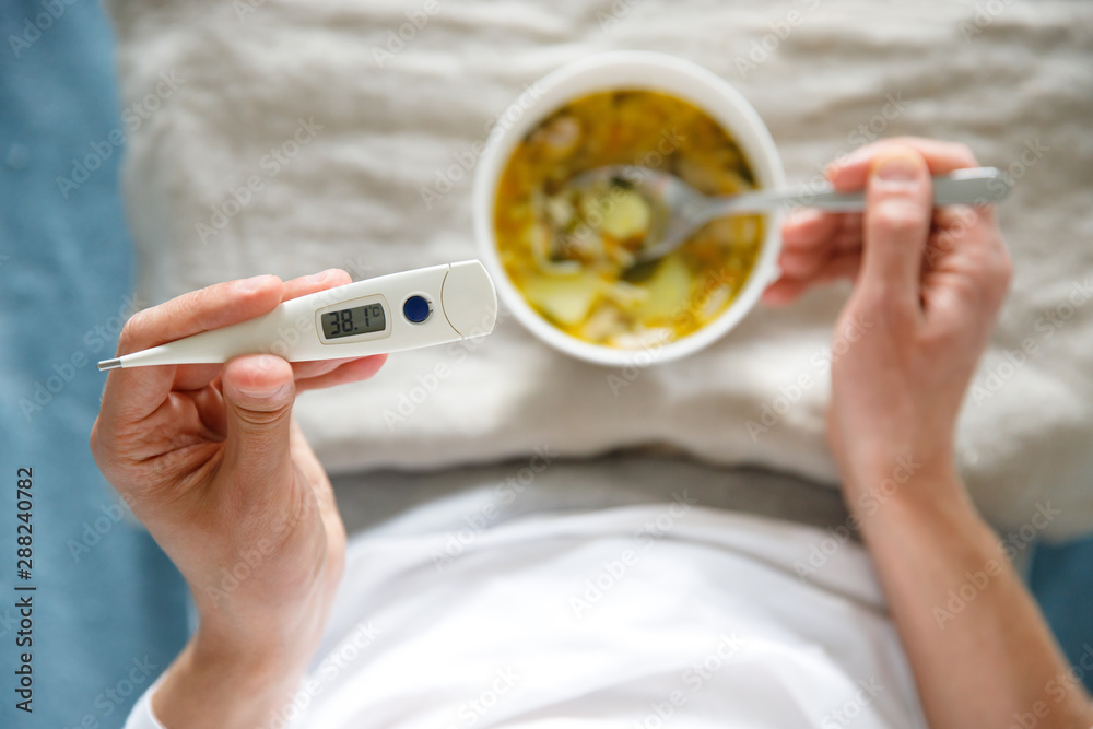 View from above diseased male eating healthy chicken soup to fight the flu sickness, holding digital thermometer in hand, soft focus.  - obrazy, fototapety, plakaty 