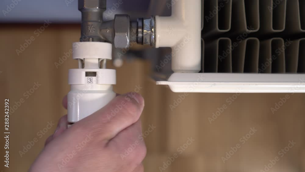 The hand turns the thermostat of a white heating radiator to reduce the room heating temperature. Blue walls and brown parquet The hand turns the thermostat of a white heating radiator to reduce the room heating temperature. Blue walls and brown parquet