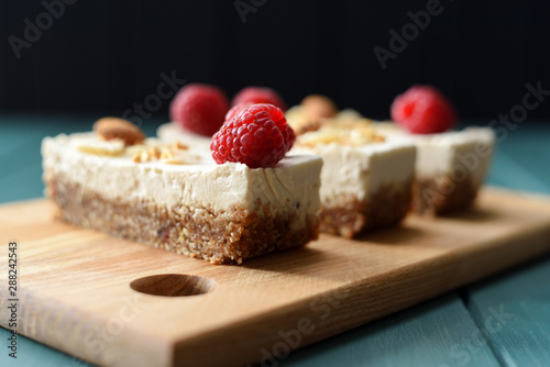 Vegan sugar free dessert. Coconut milk cheesecake bars decorated with raspberries, almonds and coconut flakes on dark background side view photo
