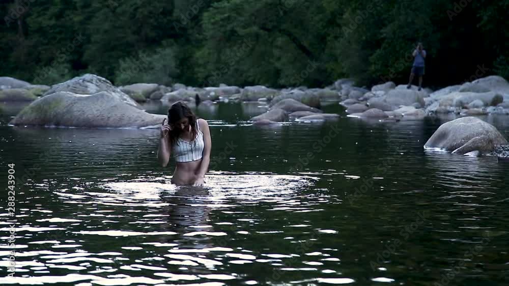 An attractive young brunette woman swimming in a pristine forest river with a creepy voyeur photographing her from afar. Stock ビデオ | Adobe Stock
