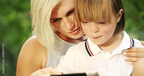 Little nice boy with Down Syndrome using educational app on tablet, spending time with mother outdoors