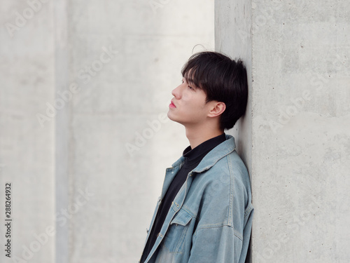 Portrait of a handsome Chinese young man in jean leaning against wall and looking away with blank expression, has nothing to do just waiting.
