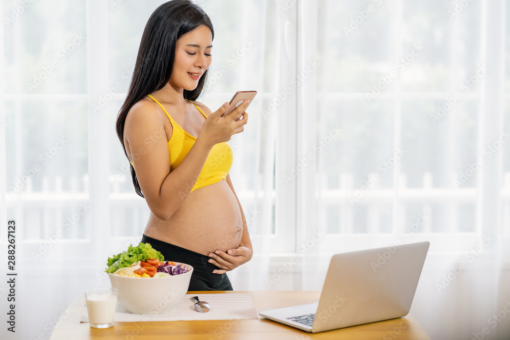 Asian Pregnant holds hands on belly and using the smart mobile phone and working with technology laptop when breakfast with Vegetable and milk on the table,preparing to be motherhood with good healthy Asian Pregnant holds hands on belly and using the smart mobile phone and working with technology laptop when breakfast with Vegetable and milk on the table,preparing to be motherhood with good healthy