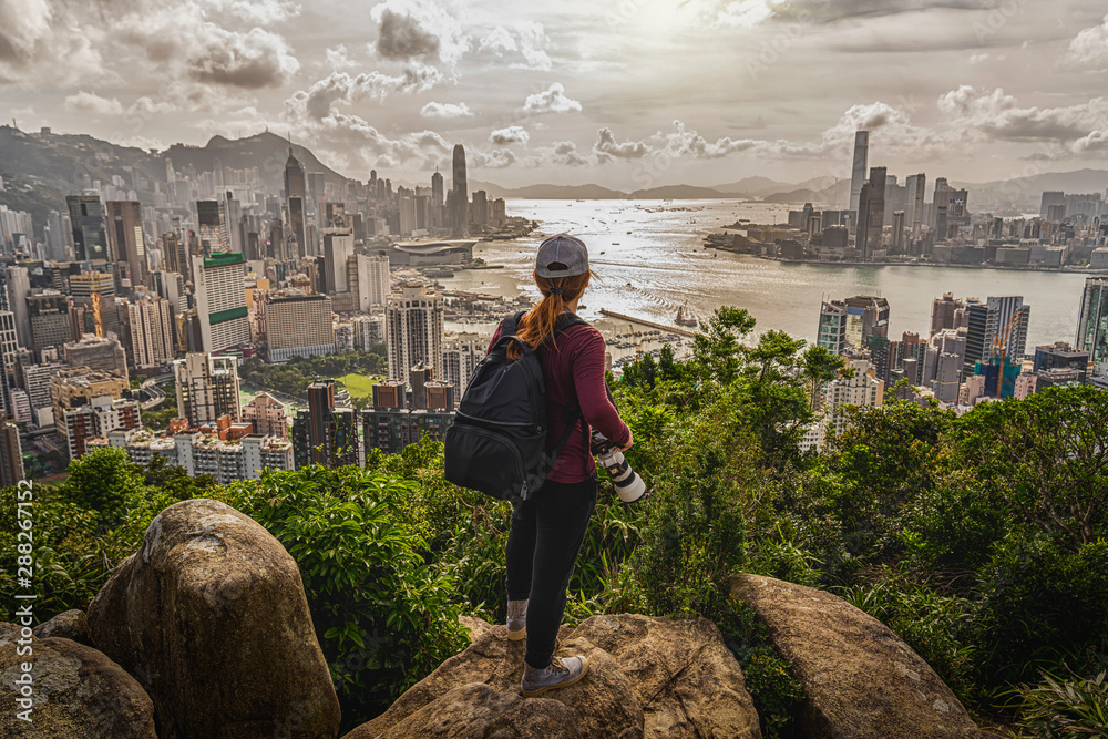 Back side of asia woman photographer standing and shooting after climbing the peak of hong kong mountain and looking hong kong and kowlloon cityscape at sunset time,adventure and travel concept