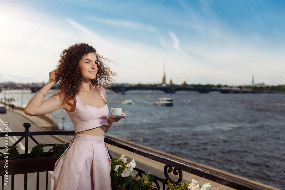 Young beautiful girl with curly hair in a pink dress is standing on the balcony and drinking coffee. Happy woman smiling and looking at the Neva River in St. ..Petersburg Young beautiful girl with curly hair in a pink dress is standing on the balcony and drinking coffee. Happy woman smiling and looking at the Neva River in St. ..Petersburg