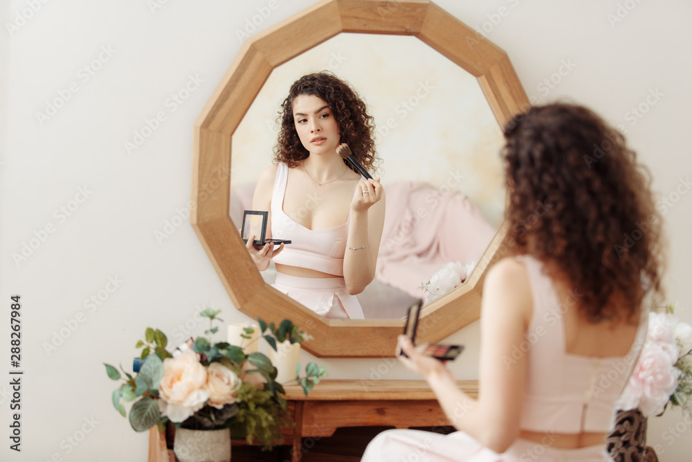 Young happy girl with curly hair does makeup in front of a vintage mirror. A beautiful woman in a pink dress paints her eyes. Young happy girl with curly hair does makeup in front of a vintage mirror. A beautiful woman in a pink dress paints her eyes.