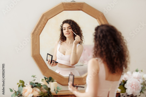 Young happy girl with curly hair does makeup in front of a vintage mirror. A beautiful woman in a pink dress paints her eyes.
