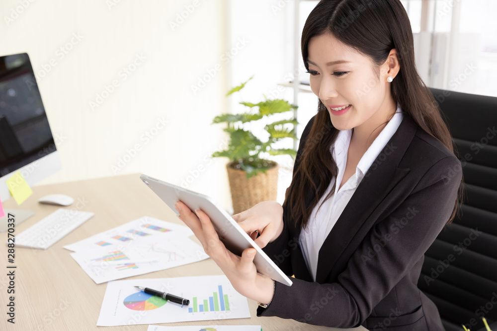 Happy Attractive businesswoman in black suit using a digital tablet while sit in desk at office building with natural light. Business and technology Happy Attractive businesswoman in black suit using a digital tablet while sit in desk at office building with natural light. Business and technology