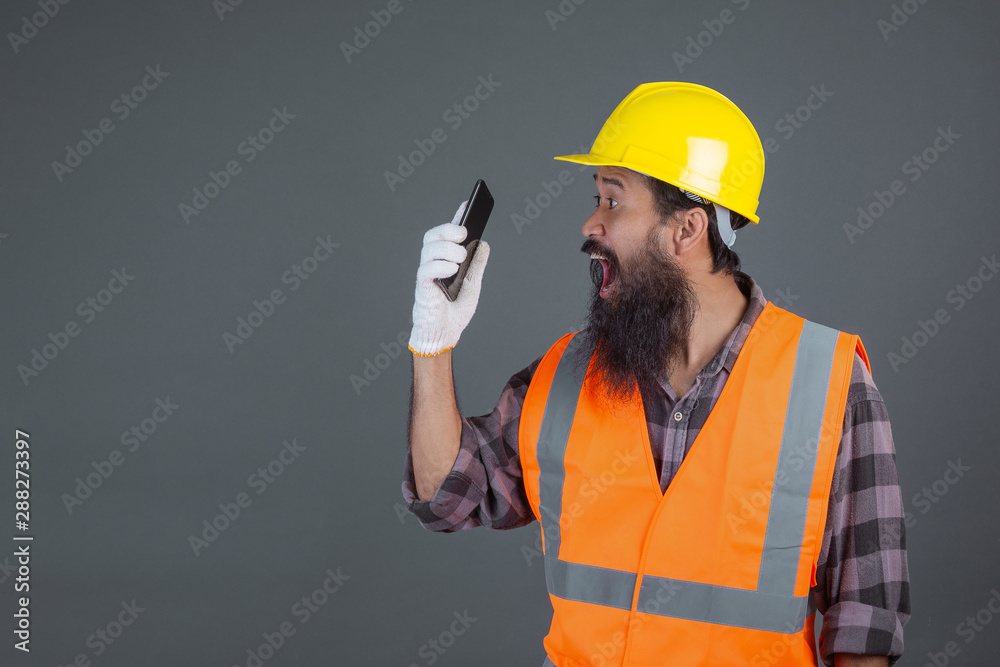 An engineering man wearing a yellow helmet holding a phone on a gray background.
