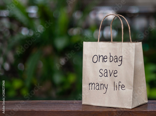 Brown recyclable paper shopping bag Placed on a wooden table, the backdroup is a green tree. For the background image used to campaign, do not use plastic bags.