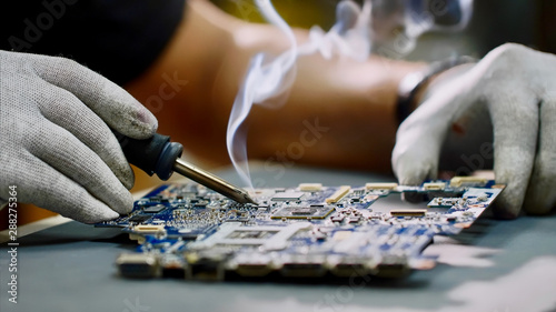 Technician engineer in workshop. Repairman in gloves is soldering circuit board of electronic device on the table, hands close up. He takes tin with a soldering iron and puts it on microcircuit. photo
