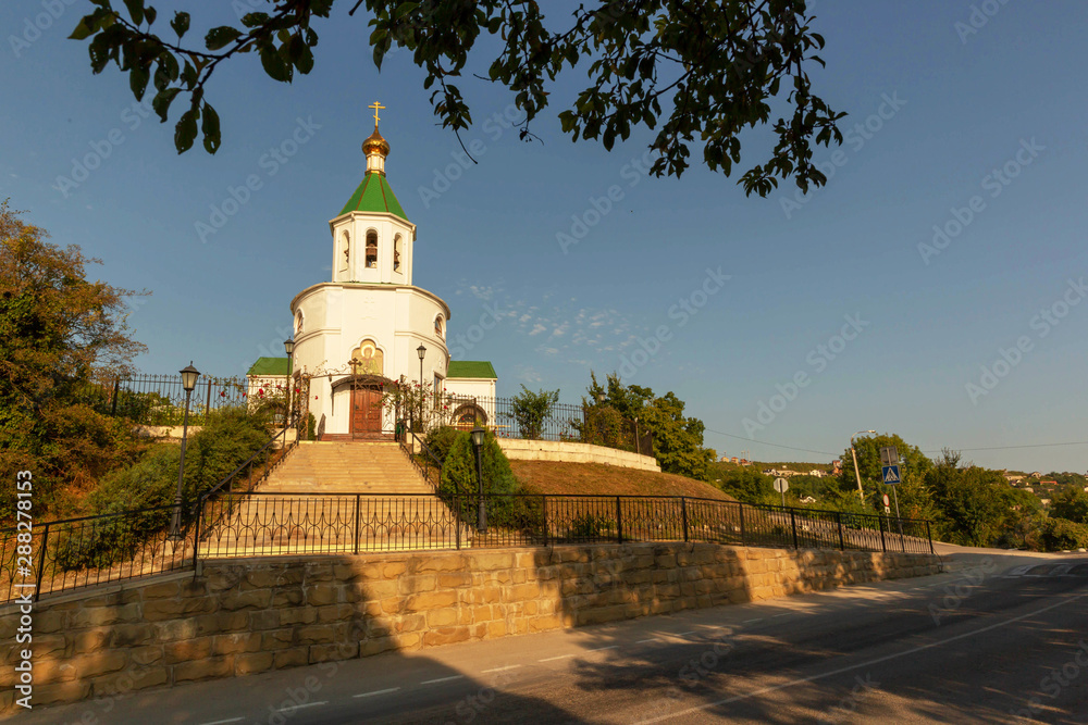 A stone white Orthodox Church with green and gold domes stands by the road.