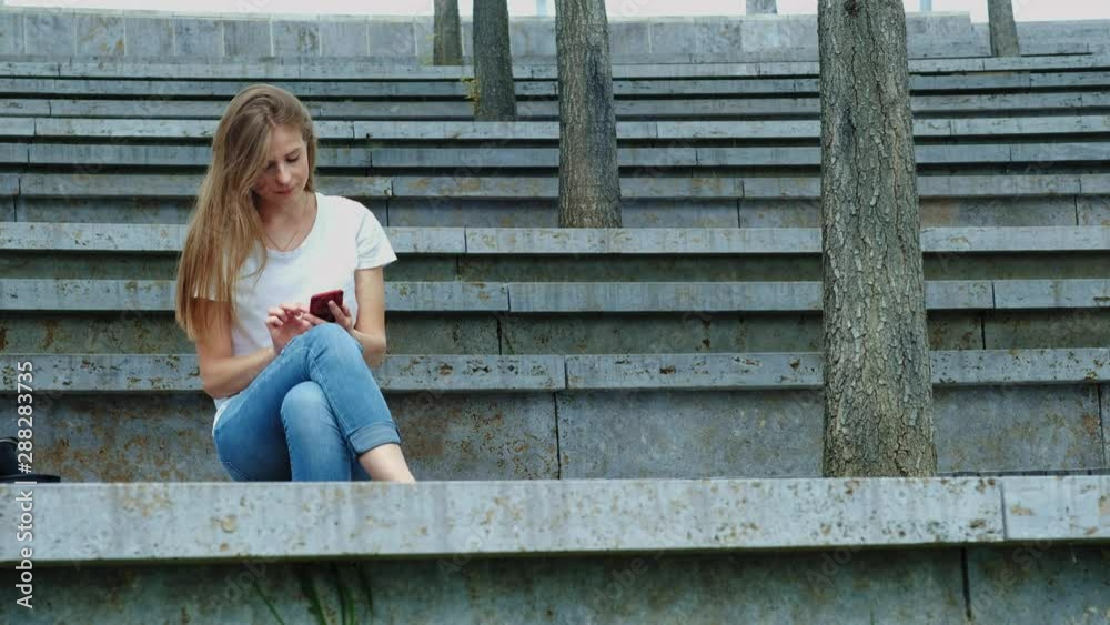 Young beautiful girl in a park holds a smartphone in her hands, writes a message. Leafing through photos on the phone, chatting in social networks with friends. Sits on concrete. Young beautiful girl in a park holds a smartphone in her hands, writes a message. Leafing through photos on the phone, chatting in social networks with friends. Sits on concrete.