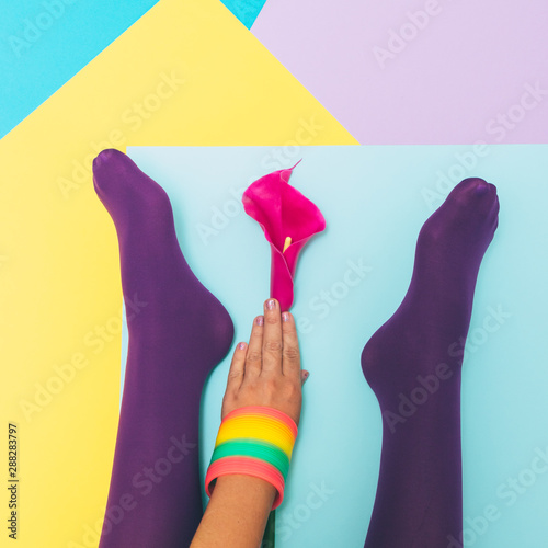 Hand with rainbow accessories touches to lily flower and legs in purple tights on a geometric blue, yellow, pink backgrounds.