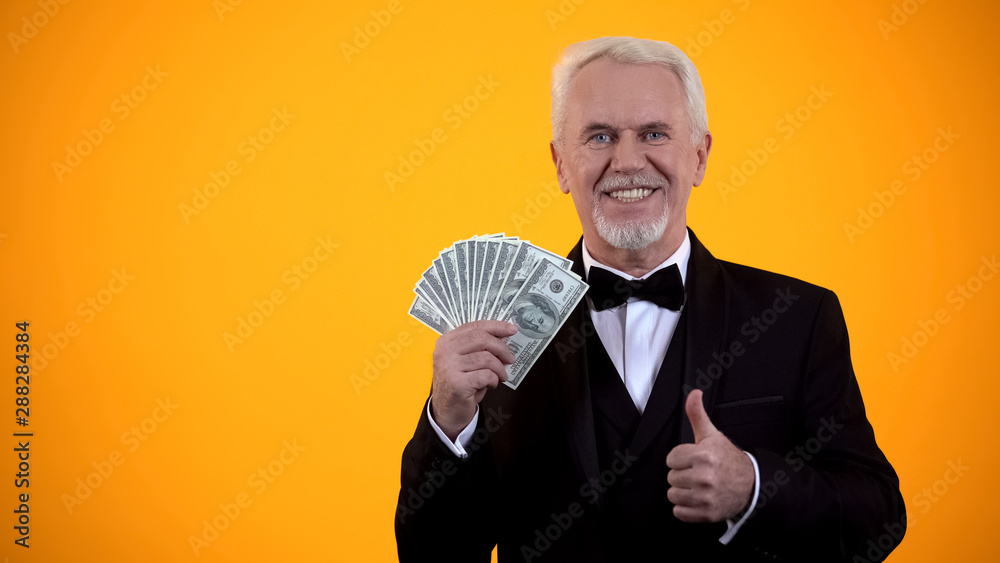 Happy old man in suit holding dollars and showing thumbs-up, business income Happy old man in suit holding dollars and showing thumbs-up, business income