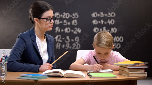 Strict teacher with ruler watching schoolboy doing his homework, control