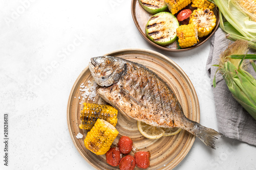 From above grilled fish with lemon served with different vegetables on dishes over white background