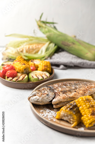 From above grilled fish with lemon served with different vegetables on dishes over white background
