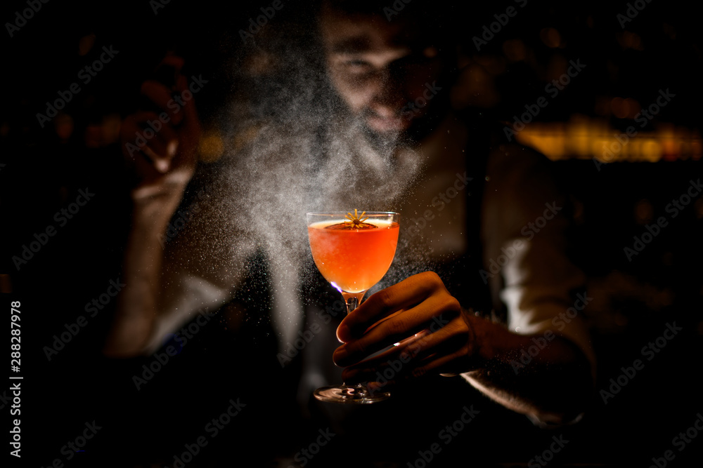 Professional bartender serving a cocktail with a lemon slice and a little yellow flower spraying on it in the dark - obrazy, fototapety, plakaty 