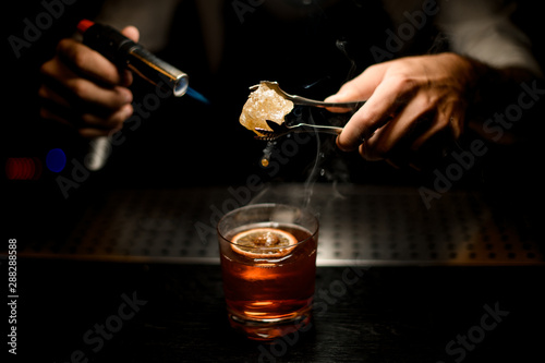 Professional bartender serving a brown cocktail melting caramel with a burner above the lemon slice in the dark