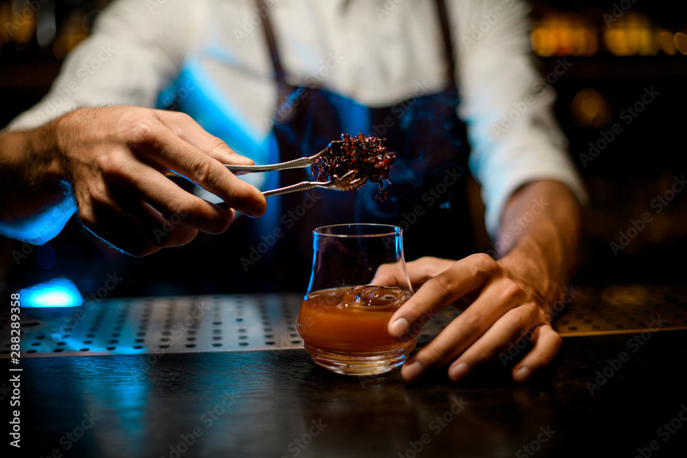 Professional bartender adding chilled melting caramel with twezzers to the cocktail glass with ice cubes under blue lights