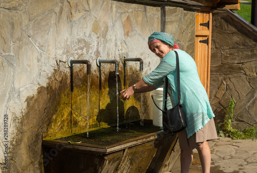 Elderly smiling woman washing an apple under water tap on summer sunny day