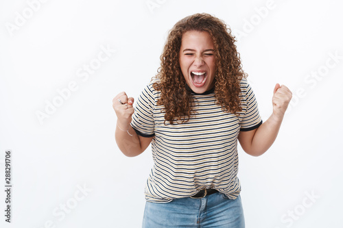 Energized confident curly-haired female showing girl power yelling out loud gather will power fists closed eyes express courage fighting stereotypes break free body-positive movement, white wall photo