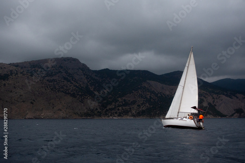 Side view of white sail boat on the grey calm water of sea or ocean during cloudy, windy and grey day with yacht team on board and rocks with green landscape on the background