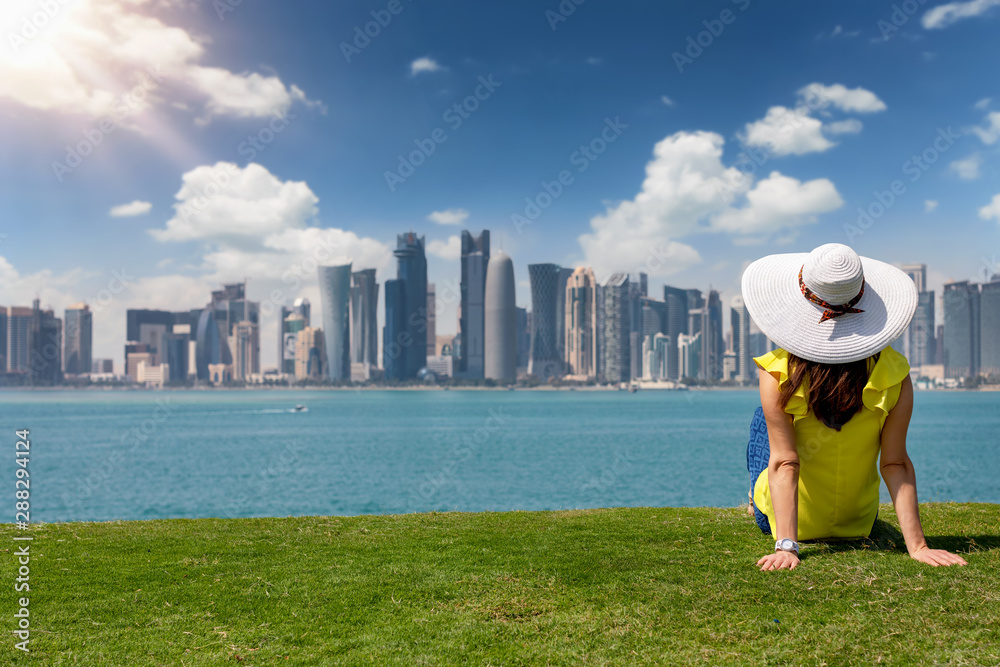 Touristin mit Sonnenhut sitzt auf einem Rasen und betrachtet die Skyline von Doha, Katar, an einem sonnigen Tag