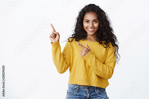 Confident good-looking african-american girl with curly hairstyle, look camera self-assured and sassy smiling, pointing upper left corner, introduce amazing product, give recommendations